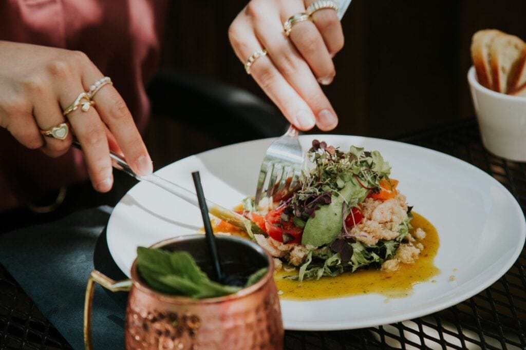 A close-up of hands cutting into a fresh plated dish with microgreens, tomatoes, avocado and prawns at Beatniks Bistro in Fort Langley, with a copper Moscow Mule mug in the foreground.