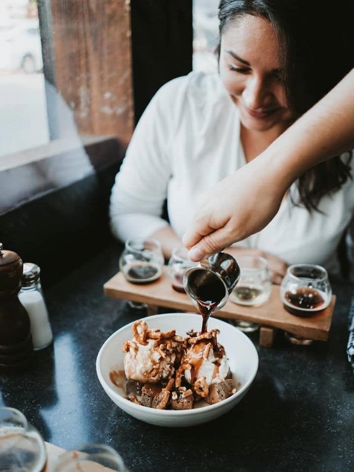 A person pours syrup over a bowl of ice cream and toppings while a woman smiles at the table in a restaurant setting.