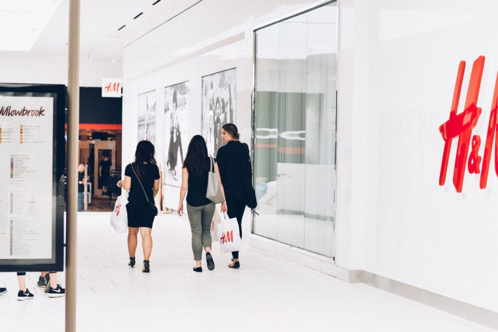 Three women walking through Willowbrook Shopping Centre in Langley, BC, carrying shopping bags past an H&M storefront.