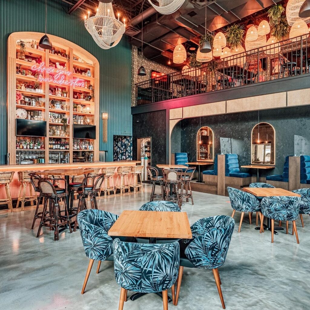 Interior of Haven Kitchen + Bar in Langley, featuring tropical print seating, a fully stocked bar with a neon "choose your own adventure" sign, woven pendant lights, and a mezzanine level above.